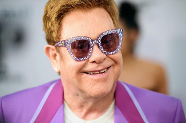 Elton John, sonriente, usando un traje  rosa y lentes con armazón lleno de estrellas.